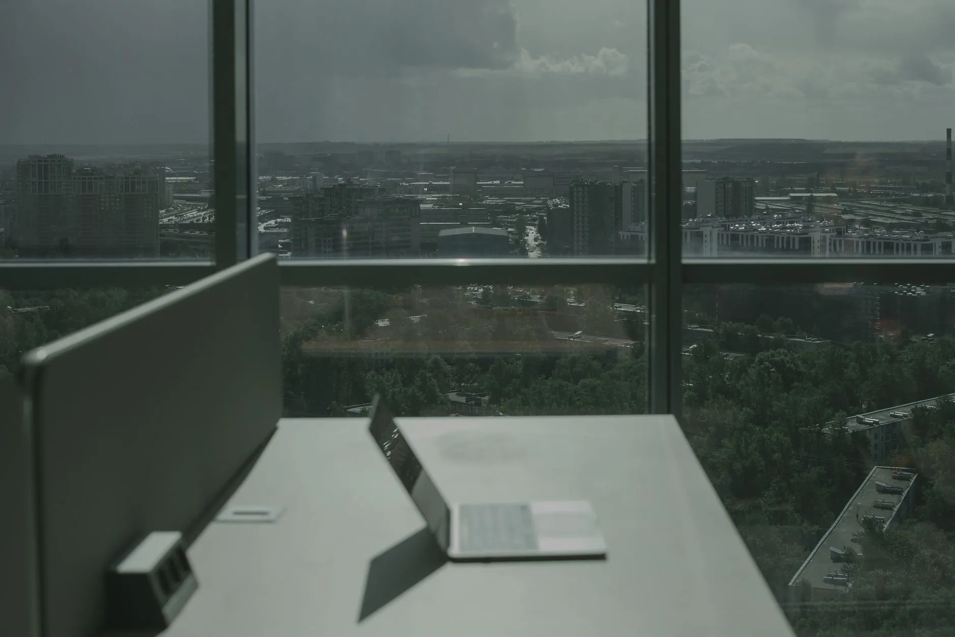 An empty desk with an open laptop beside a tall office window over a grey city