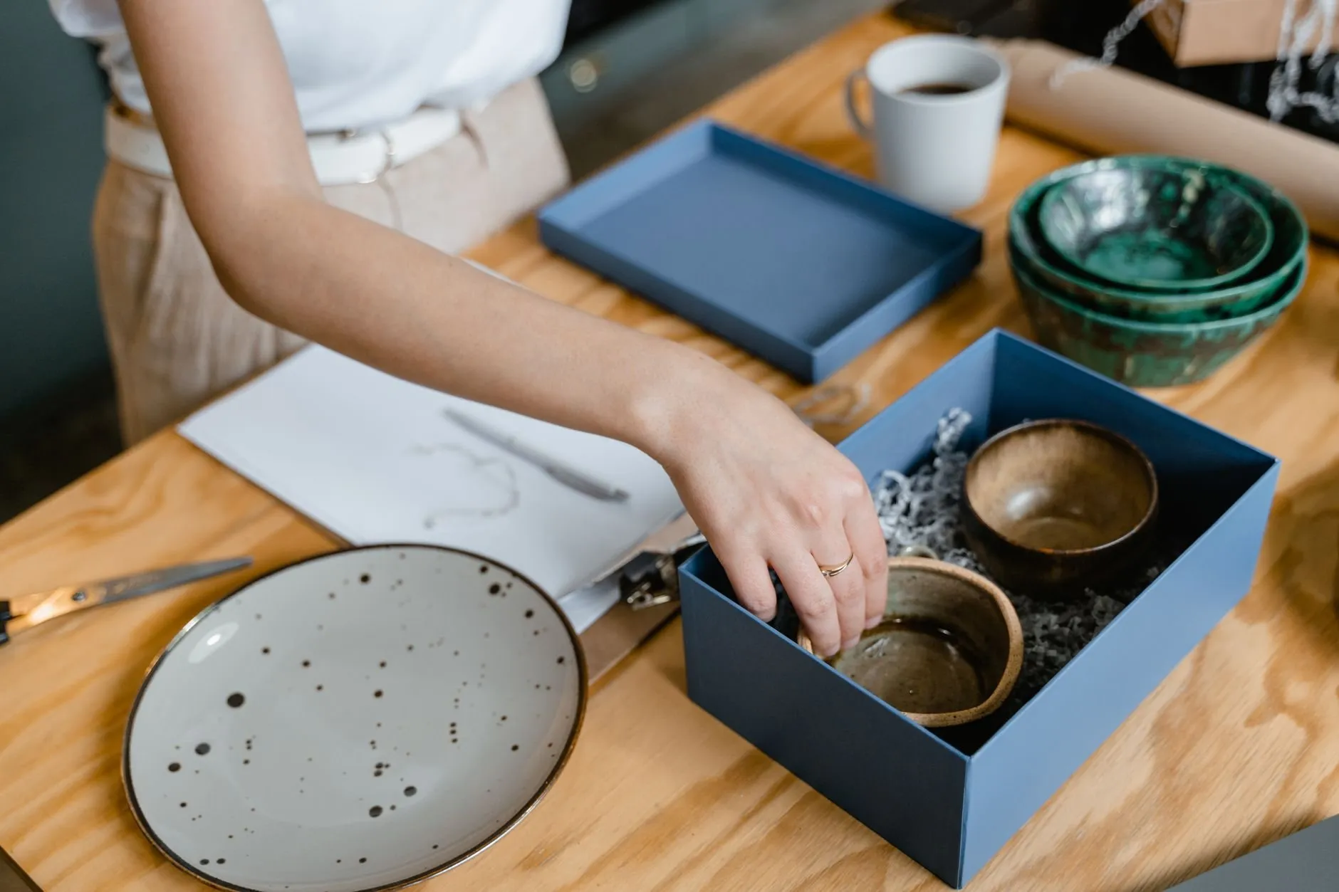 Hands placing a stoneware bowl into a blue gift box on a wooden packing table