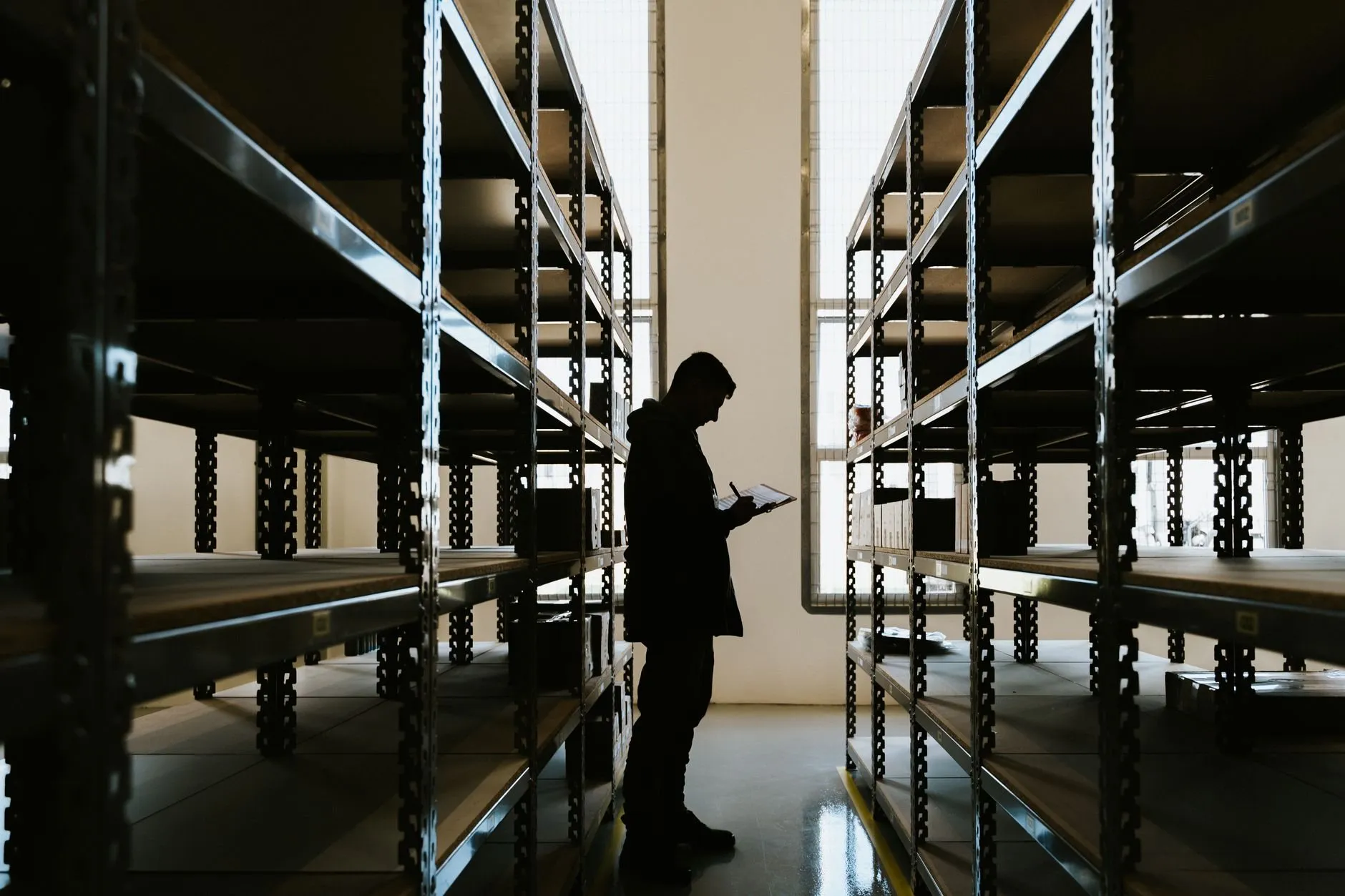 A silhouetted person checking a clipboard between rows of metal warehouse shelving
